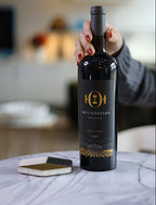 A hand with red nail polish holds a bottle of wine from Oak & Vineyard next to a small notebook on a white marble table in a modern, softly blurred interior setting.